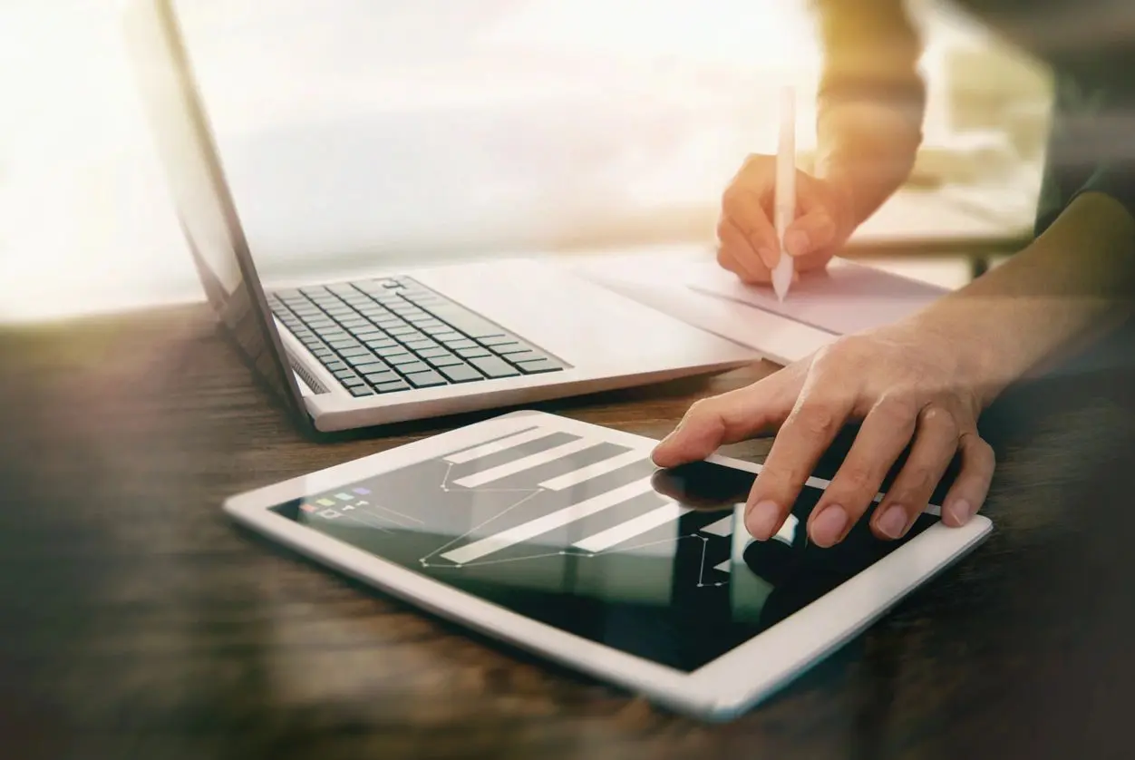 Person using a tablet and writing notes beside a laptop at a sunny desk.