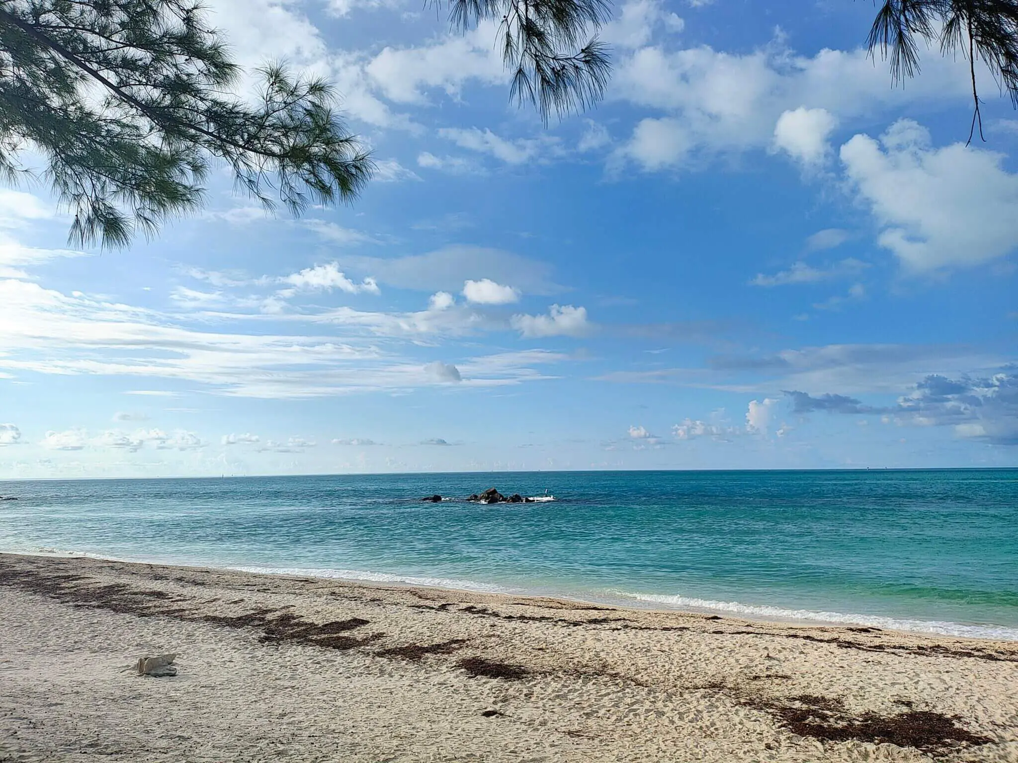 Sunny beach with clear blue water and scattered clouds.